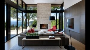 Modern living room with floor-to-ceiling windows, stone fireplace, grey sofa, red chairs, and scenic outdoor view.